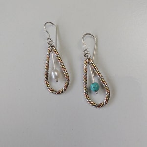 May include: A pair of silver, copper and gold teardrop earrings with a white pearl and a turquoise stone hanging inside each teardrop.