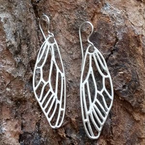 May include: A pair of silver earrings shaped like dragonfly wings. The wings are delicate and intricate, with a detailed vein pattern.