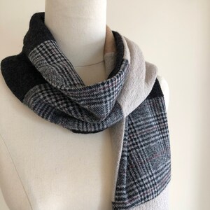 May include: A patterned scarf draped on a mannequin. The scarf combines sections of black, grey, and beige fabric. A section of the scarf has a classic black and grey plaid pattern with red accents. The scarf is wrapped around the mannequin's neck.