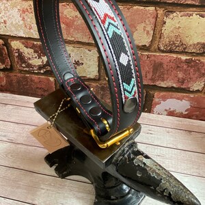 May include: Black leather dog collar with a red stitched edge and a colorful geometric pattern. The collar is on a black metal anvil.