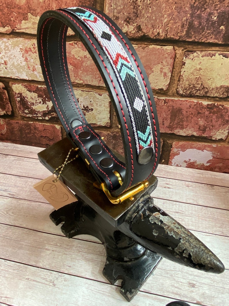 May include: Black leather dog collar with a red stitched edge and a colorful geometric pattern. The collar is on a black metal anvil.