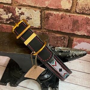 May include: Black leather dog collar with a red and white geometric pattern and gold hardware. The collar is on a black anvil.