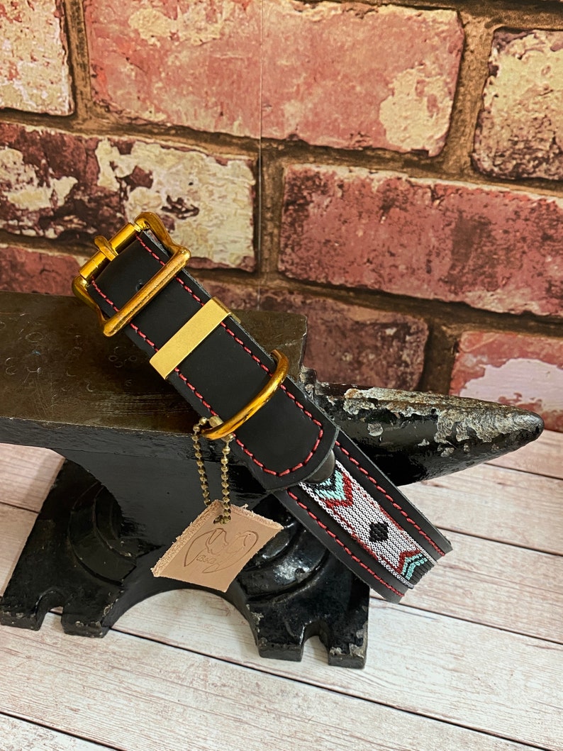 May include: Black leather dog collar with a red and white geometric pattern and gold hardware. The collar is on a black anvil.