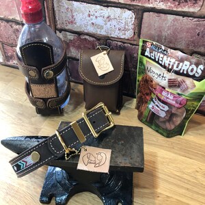 May include: A brown leather dog collar with a gold buckle and a beaded design. The collar is on a black metal anvil with a brown leather dog tag with a paw print design. There is a brown leather water bottle holder with a red lid and a brown leather pouch with a gold tag. A bag of dog treats is in the background.