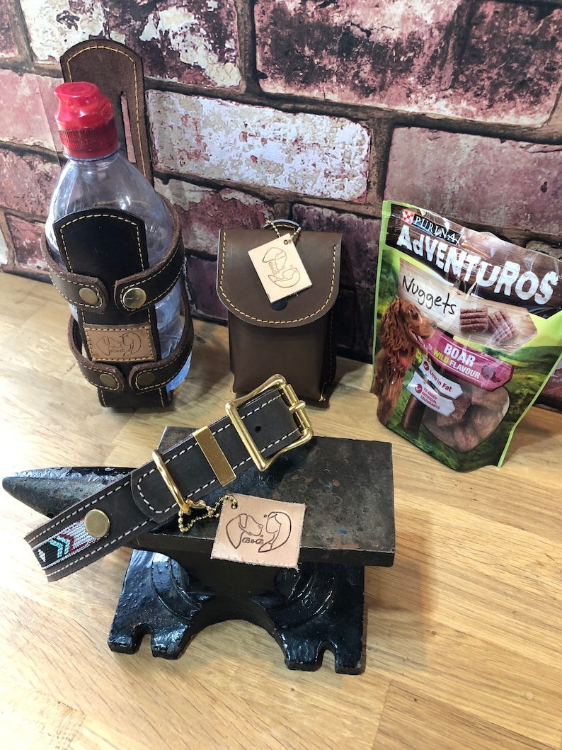 May include: A brown leather dog collar with a gold buckle and a beaded design. The collar is on a black metal anvil with a brown leather dog tag with a paw print design. There is a brown leather water bottle holder with a red lid and a brown leather pouch with a gold tag. A bag of dog treats is in the background.