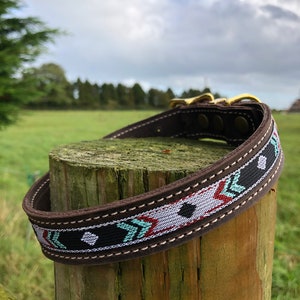 May include: A brown leather dog collar with a colourful geometric pattern. The collar has a gold metal buckle and is wrapped around a wooden post.