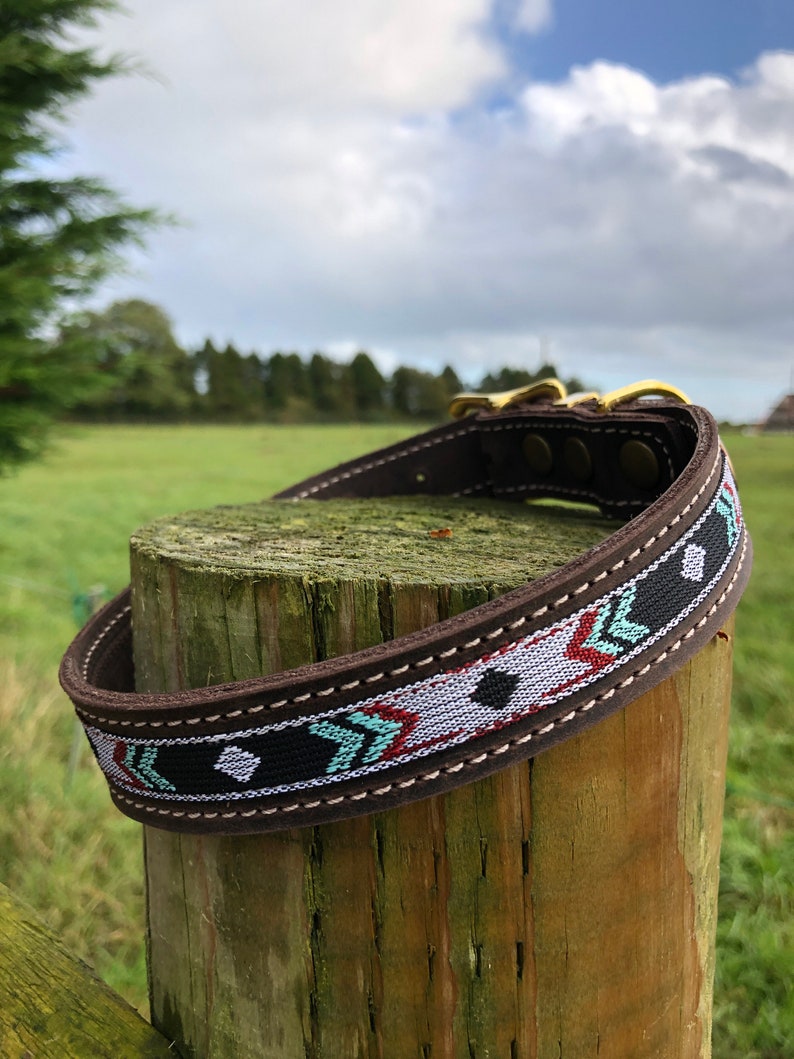 May include: A brown leather dog collar with a colourful geometric pattern. The collar has a gold metal buckle and is wrapped around a wooden post.