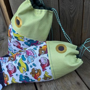 May include: Two drawstring bags shaped like fish. The fish-shaped bags have a light green head with a yellow eye and a body patterned with colorful cartoon fish. The drawstring is green and white. The bags are on a wooden surface.