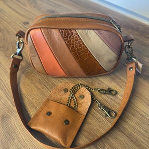 Peut inclure: Un sac banane en cuir marron avec un motif rayé multicolore et une petite pochette assortie. Le sac banane a une fermeture éclair et une bandoulière amovible. La pochette a une chaîne et un fermoir.