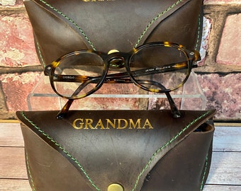 May include: Two brown leather eyeglass cases with green stitching. The top case has the word "GRANDPA" embossed in gold lettering. The bottom case has the word "GRANDMA" embossed in gold lettering.