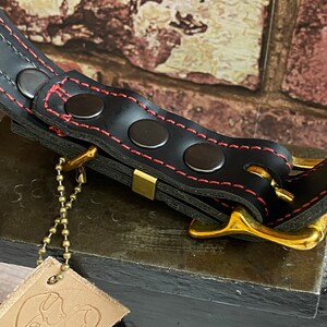 May include: Black leather dog collar with red stitching and brass hardware. The collar has a chain attached to a leather tag with a dog illustration and the text "Bell".