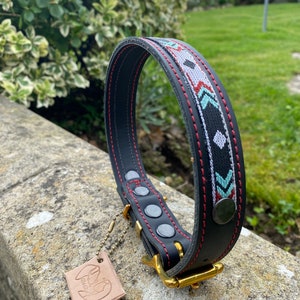 May include: Black leather dog collar with red stitching and a colorful geometric pattern. The collar has a gold metal buckle and a leather tag with the word "Lucky" on it.