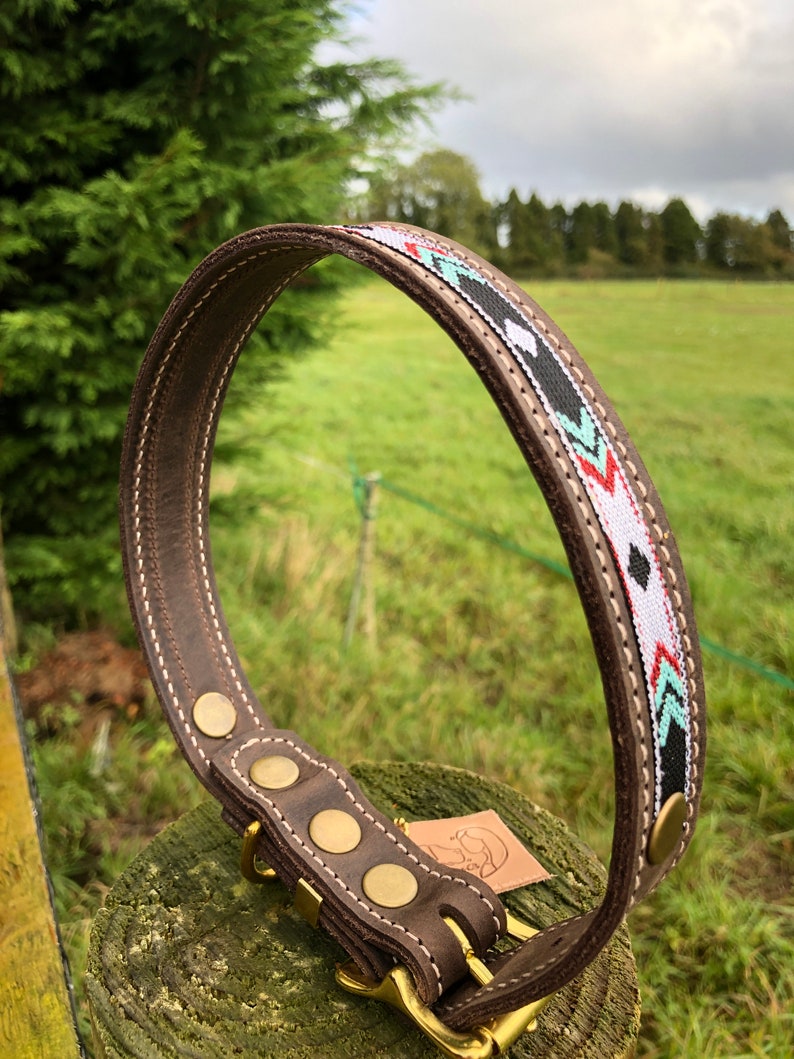 May include: Brown leather dog collar with a white, black, red and green geometric pattern. The collar has brass hardware and four brass rivets.
