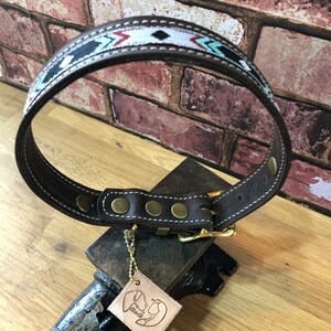 May include: Brown leather dog collar with a white, black and turquoise geometric pattern. The collar has a brass buckle and five brass rivets. The collar is on a metal anvil with a leather tag that says "Pawsitive".