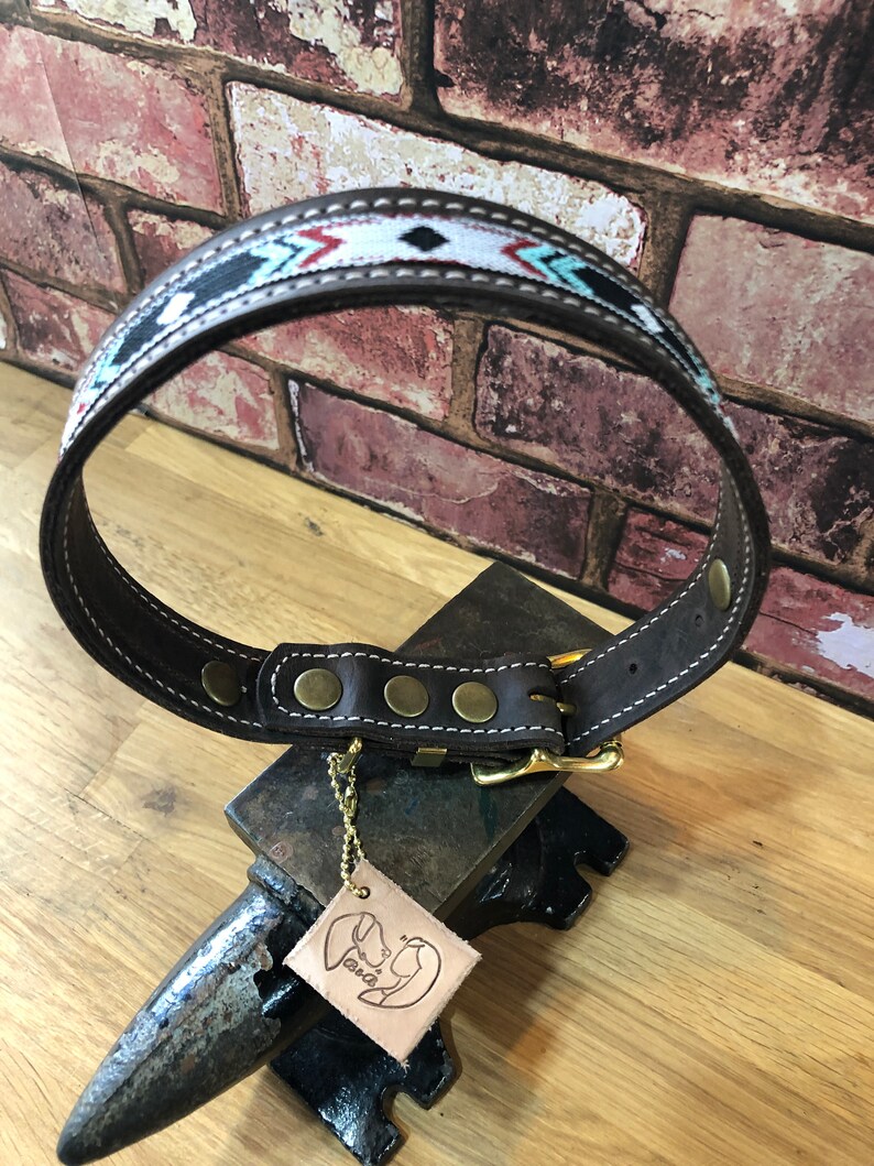 May include: Brown leather dog collar with a white, black and turquoise geometric pattern. The collar has a brass buckle and five brass rivets. The collar is on a metal anvil with a leather tag that says "Pawsitive".
