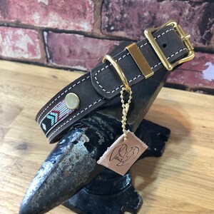 May include: A brown leather dog collar with a gold buckle and a decorative woven strap. The collar is on a black metal anvil with a leather tag that says "B&D".