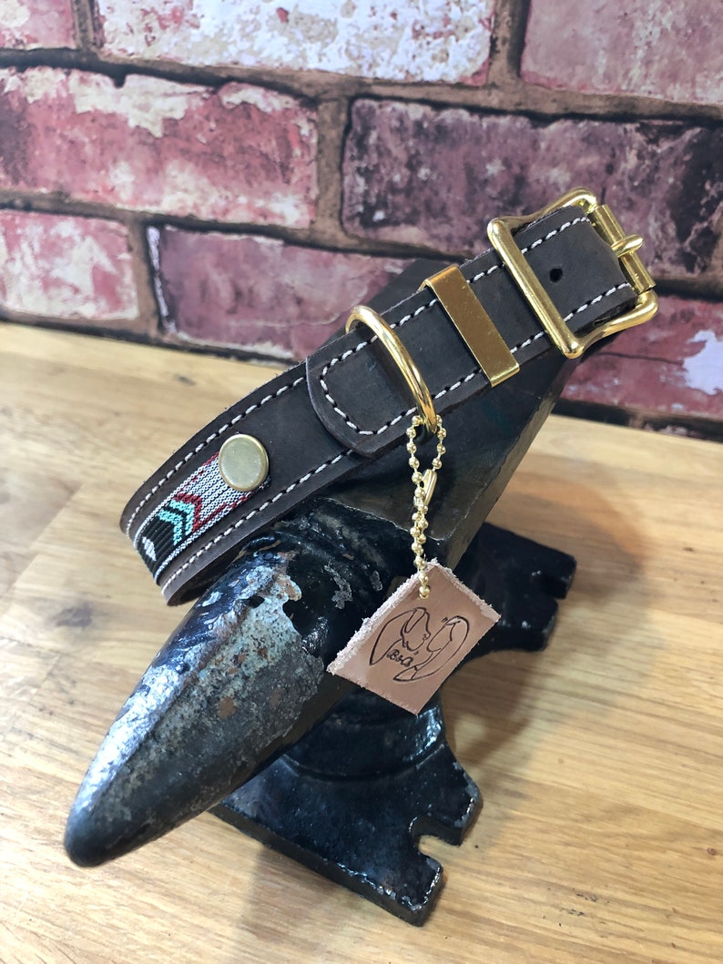 May include: A brown leather dog collar with a gold buckle and a decorative woven strap. The collar is on a black metal anvil with a leather tag that says "B&D".