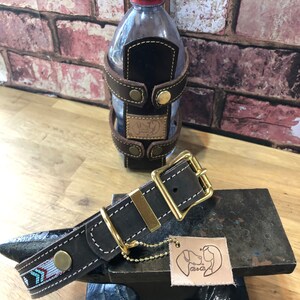May include: A brown leather dog collar with a gold buckle and a small chain attached. The collar has a decorative pattern of blue, white and red arrows. The collar is sitting on a black metal surface.