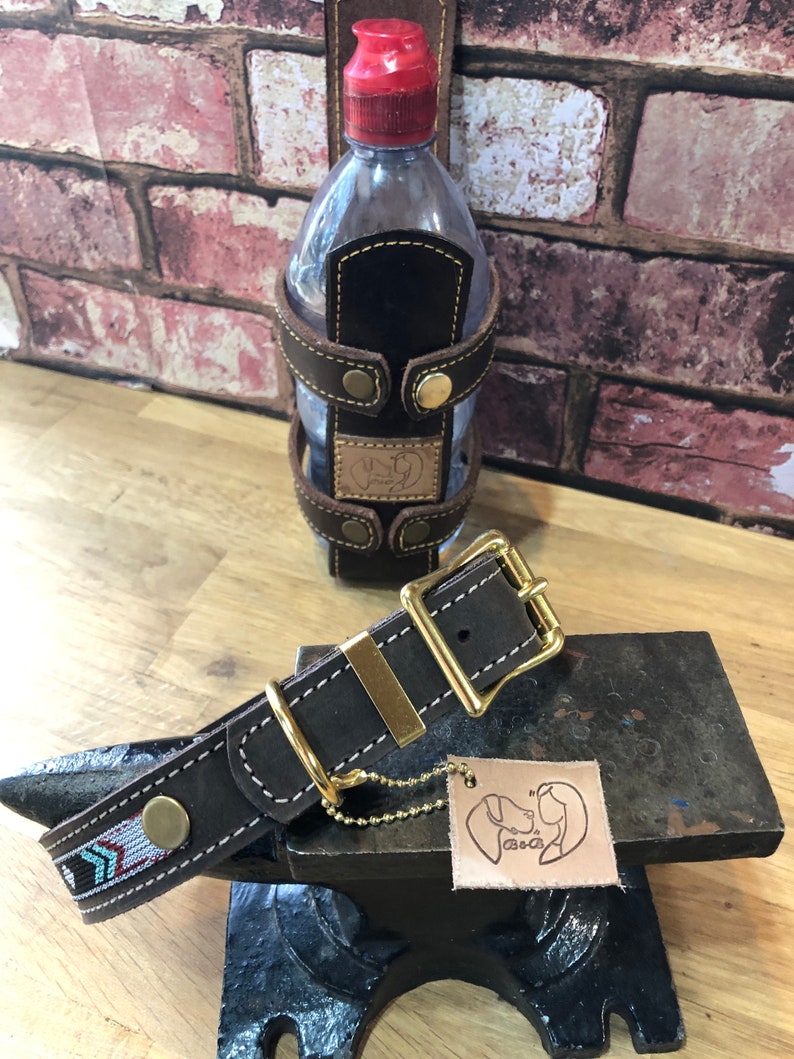 May include: A brown leather dog collar with a gold buckle and a small chain attached. The collar has a decorative pattern of blue, white and red arrows. The collar is sitting on a black metal surface.