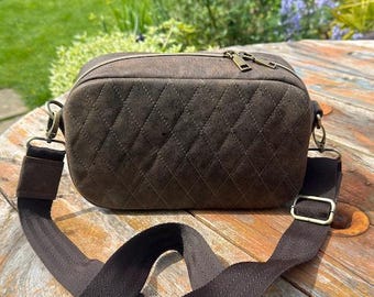 May include: A brown, quilted crossbody bag with a rectangular shape. The bag features a zippered top, a long adjustable strap, and gold-colored hardware. The bag is made of a soft, suede-like material.