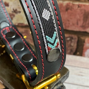 May include: A black leather dog collar with a red stitched edge and a colorful woven pattern. The collar has a gold metal buckle and a silver metal rivet.