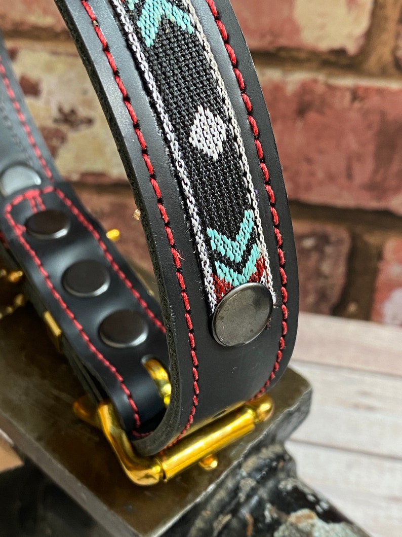 May include: A black leather dog collar with a red stitched edge and a colorful woven pattern. The collar has a gold metal buckle and a silver metal rivet.