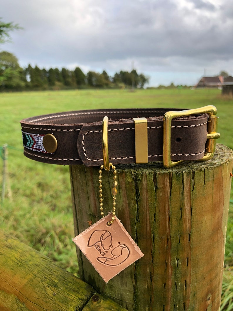 May include: Brown leather dog collar with a brass buckle and a decorative woven strip. The collar is attached to a wooden post with a leather tag that says "B&B".