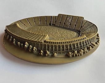 May include: A bronze stadium model with detailed features. The oval design includes tiered seating, a central field, and a raised section with a crest. The model has a textured surface and sits on a flat base.