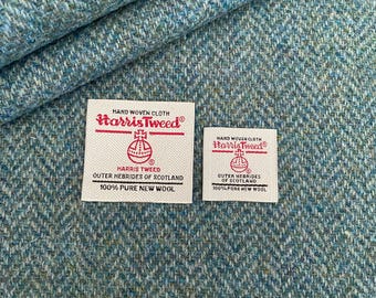 Peut inclure: Deux étiquettes blanches avec le texte "HAND WOVEN CLOTH Harris Tweed®" et le logo Harris Tweed Orb. Le texte sous le logo indique "HARRIS TWEED OUTER HEBRIDES OF SCOTLAND 100% PURE NEW WOOL". Les étiquettes sont sur un tissu en tweed bleu et vert à chevrons.