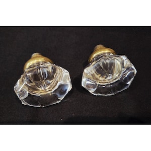 May include: Two clear glass cabinet knobs with a faceted design and brass-colored metal bases. The knobs are set against a dark background, highlighting their clarity and shape. These vintage-style knobs are suitable for furniture restoration or adding a touch of elegance.