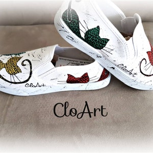 May include: A pair of white canvas slip-on shoes with hand-painted cat designs. The shoes feature a yellow, green, and red cat on each shoe. The shoes are signed "CloArt" on the side.