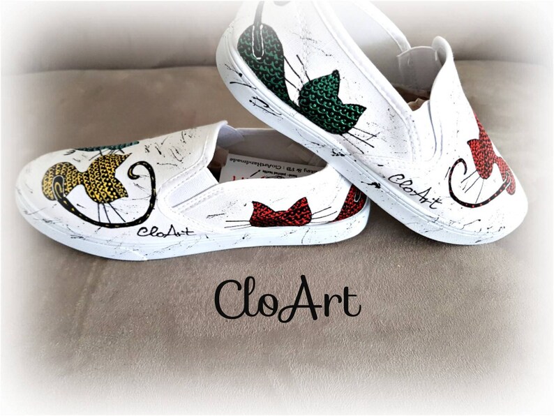 May include: A pair of white canvas slip-on shoes with hand-painted cat designs. The shoes feature a yellow, green, and red cat on each shoe. The shoes are signed "CloArt" on the side.