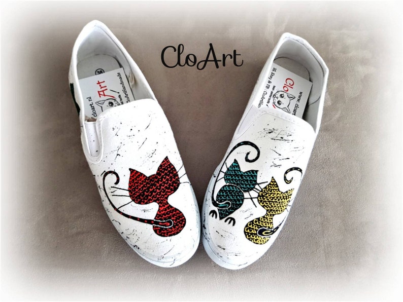 May include: A pair of white canvas shoes with hand-painted cat designs. One shoe features a red cat, the other features a green and yellow cat. The shoes have a black splatter paint effect.