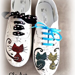 May include: A pair of white canvas shoes with hand-painted cat designs. One shoe features a red cat, the other has a blue and yellow cat. The shoes have black and white polka dot laces and a blue ribbon.