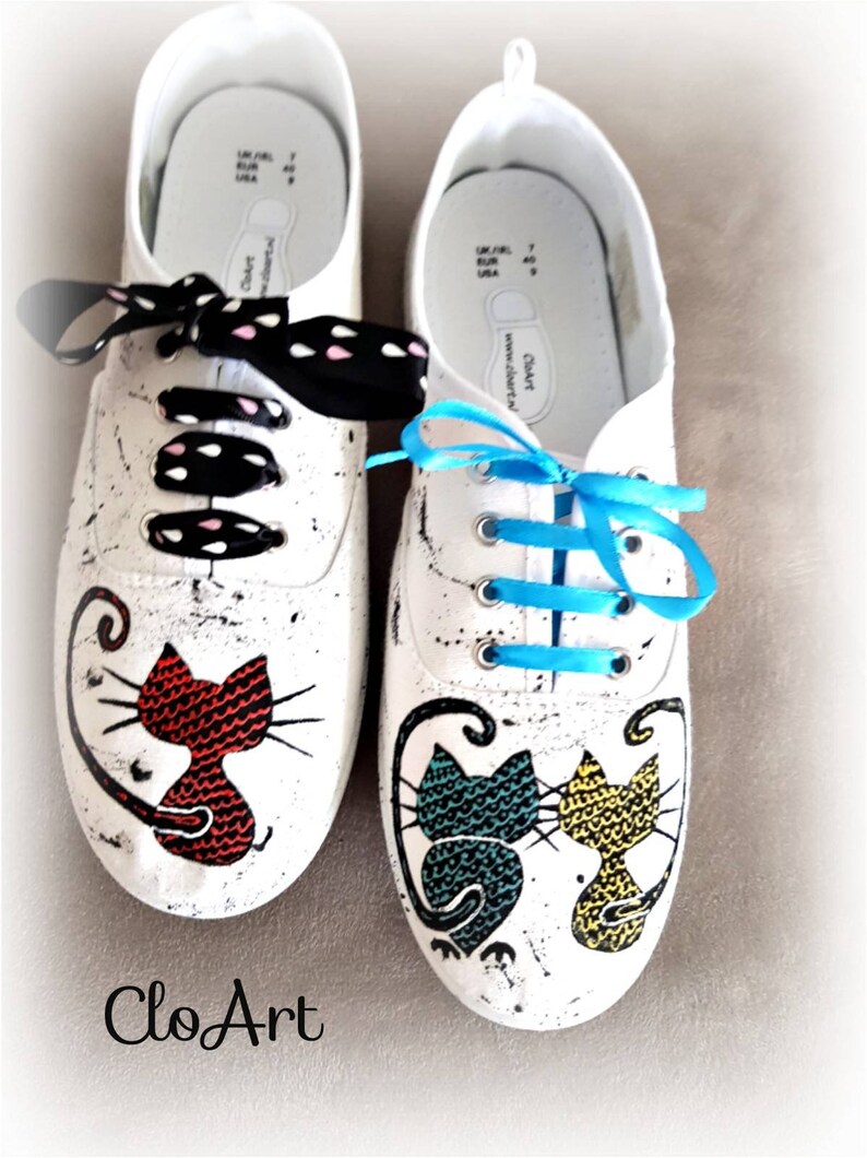 May include: A pair of white canvas shoes with hand-painted cat designs. One shoe features a red cat, the other has a blue and yellow cat. The shoes have black and white polka dot laces and a blue ribbon.