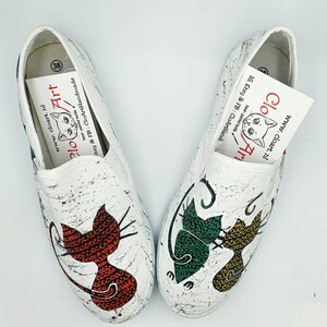 May include: A pair of white canvas slip-on shoes with hand-painted cat designs. One shoe features a red cat, the other a green and yellow cat. The shoes are decorated with black paint splatters.