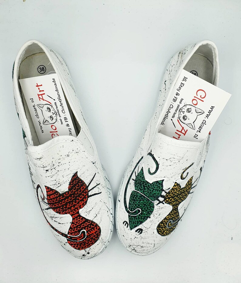 May include: A pair of white canvas slip-on shoes with hand-painted cat designs. One shoe features a red cat, the other a green and yellow cat. The shoes are decorated with black paint splatters.