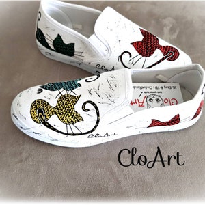May include: A pair of white canvas slip-on shoes with hand-painted cat designs. The shoes feature a black cat with green accents, a yellow cat with black and white accents, and a red cat with white accents. The shoes are decorated with the text "Clo Art" and "Clasit".