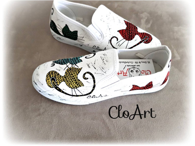 May include: A pair of white canvas slip-on shoes with hand-painted cat designs. The shoes feature a black cat with green accents, a yellow cat with black and white accents, and a red cat with white accents. The shoes are decorated with the text "Clo Art" and "Clasit".