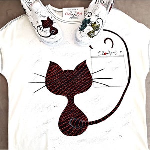 May include: White t-shirt with a black cat design with red scales. The cat has a long tail that wraps around the shirt. The shirt also has a pocket with the text "CloArt" on it. Two white sneakers with colorful designs are on top of the shirt.