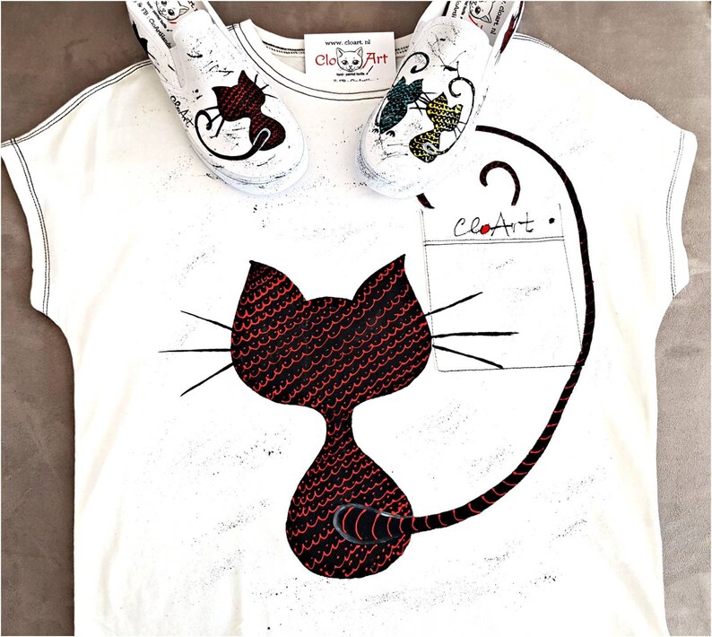 May include: White t-shirt with a black cat design with red scales. The cat has a long tail that wraps around the shirt. The shirt also has a pocket with the text "CloArt" on it. Two white sneakers with colorful designs are on top of the shirt.