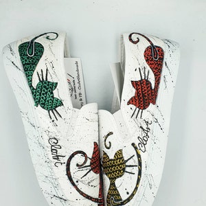 May include: A pair of white canvas slip-on shoes with hand-painted black and white splatter paint. Each shoe features a different cat design with a green, red, and yellow patterned body. The text "CloArt" is written on each shoe.