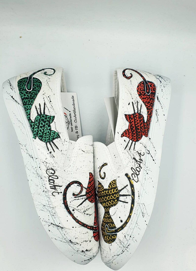 May include: A pair of white canvas slip-on shoes with hand-painted black and white splatter paint. Each shoe features a different cat design with a green, red, and yellow patterned body. The text "CloArt" is written on each shoe.