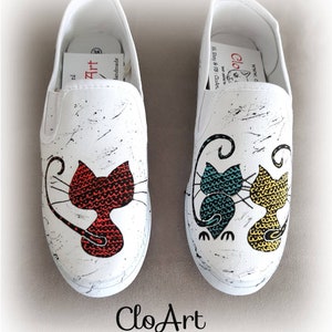 May include: A pair of white canvas slip-on shoes with hand-painted cat designs. One shoe features a red cat, while the other shoe features two cats, one teal and one yellow. The shoes have black splatter paint accents and the text "CloArt" is written on the bottom of the shoes.