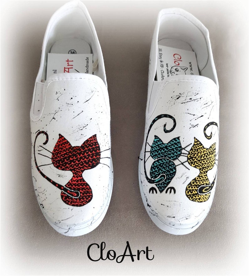 May include: A pair of white canvas slip-on shoes with hand-painted cat designs. One shoe features a red cat, while the other shoe features two cats, one teal and one yellow. The shoes have black splatter paint accents and the text "CloArt" is written on the bottom of the shoes.
