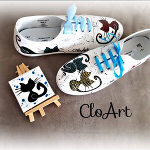 May include: White canvas shoes with hand-painted black cat designs. The shoes have blue laces and are displayed with a small easel holding a canvas with a black cat painting. The shoes are labeled "CloArt".