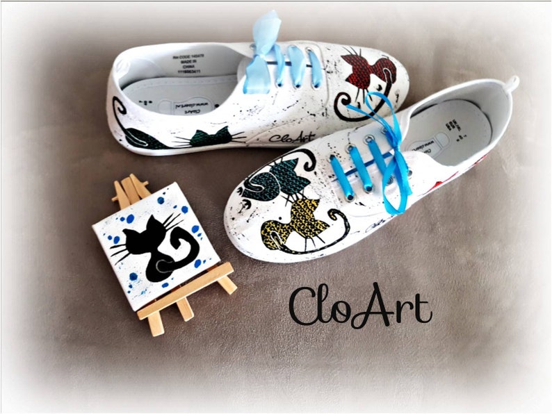 May include: White canvas shoes with hand-painted black cat designs. The shoes have blue laces and are displayed with a small easel holding a canvas with a black cat painting. The shoes are labeled "CloArt".