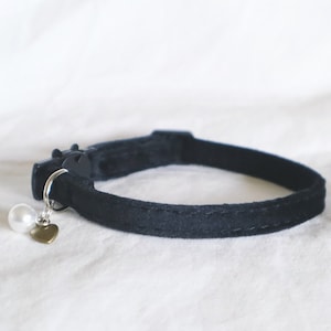 Pode incluir: Um colar de veludo preto para gato com um pingente de coração prateado e uma pérola branca.