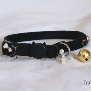 May include: Black cat collar with a black plastic cat-shaped buckle and a gold bell. The collar has small colorful beads and a silver charm.