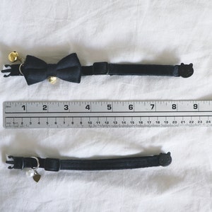May include: Two black cat collars with different charms. The top collar has a black bow tie with gold bells. The bottom collar has a small silver heart charm with a pearl.